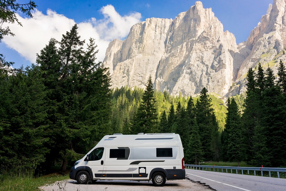 A modern camper van is parked on a scenic roadside, surrounded by lush green trees and towering rocky mountains. The clear blue sky enhances the natural landscape, providing an ideal backdrop for outdoor adventures.