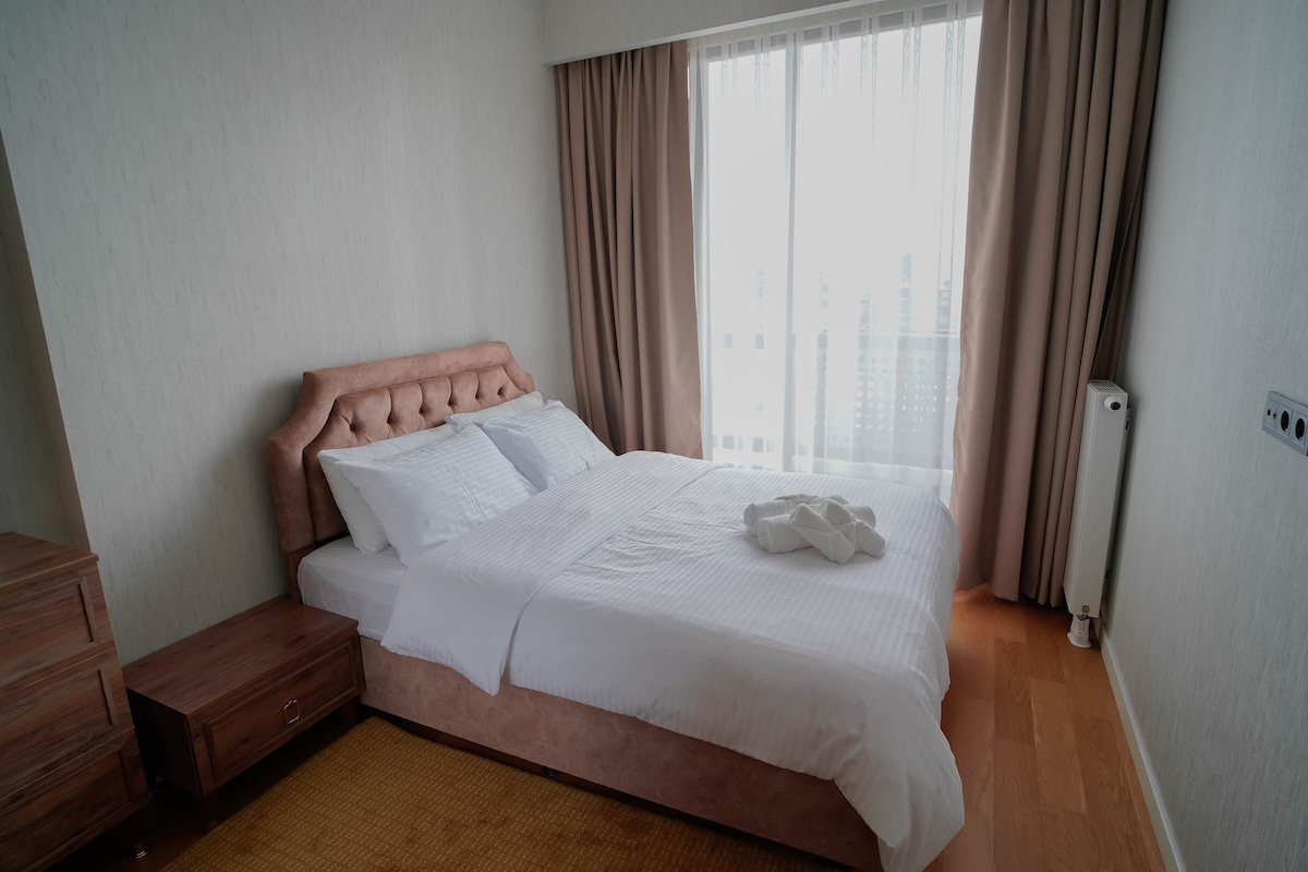 A well-lit bedroom features a spacious bed with soft white linens, complemented by a plush headboard. Natural light floods the room through large windows, with sheer curtains gently diffusing the sunlight. A wooden nightstand and a radiator are visible, enhancing the modern design.