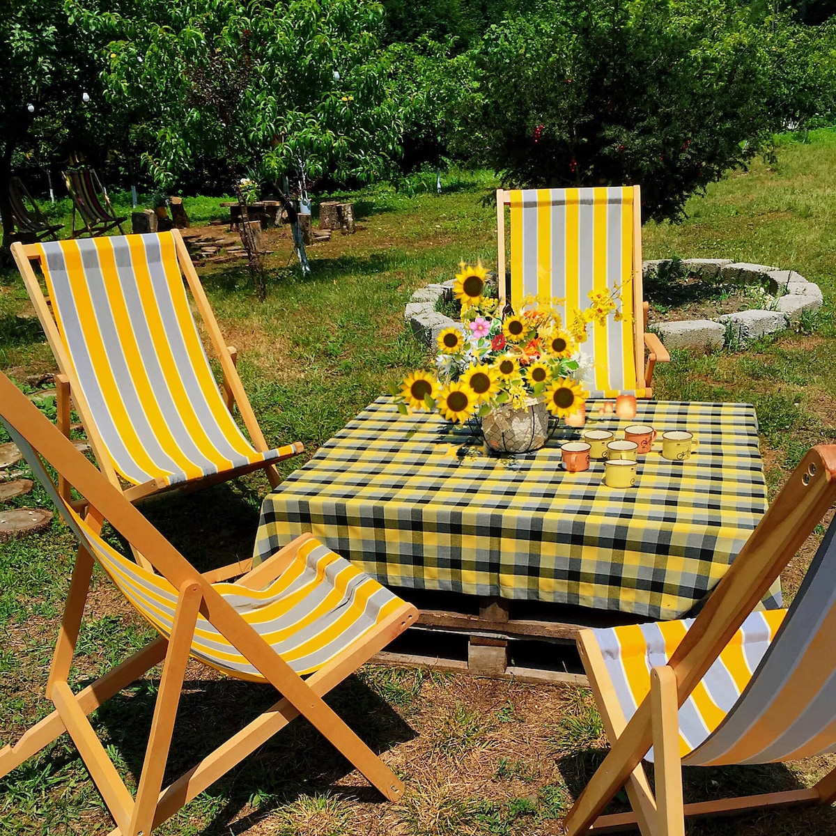 A wooden table is set outdoors, surrounded by four yellow and white striped chairs. A checkered tablecloth drapes over the table, where a vase filled with sunflowers sits amidst cups and small plates. Lush greenery surrounds the gathering area, providing a tranquil setting.