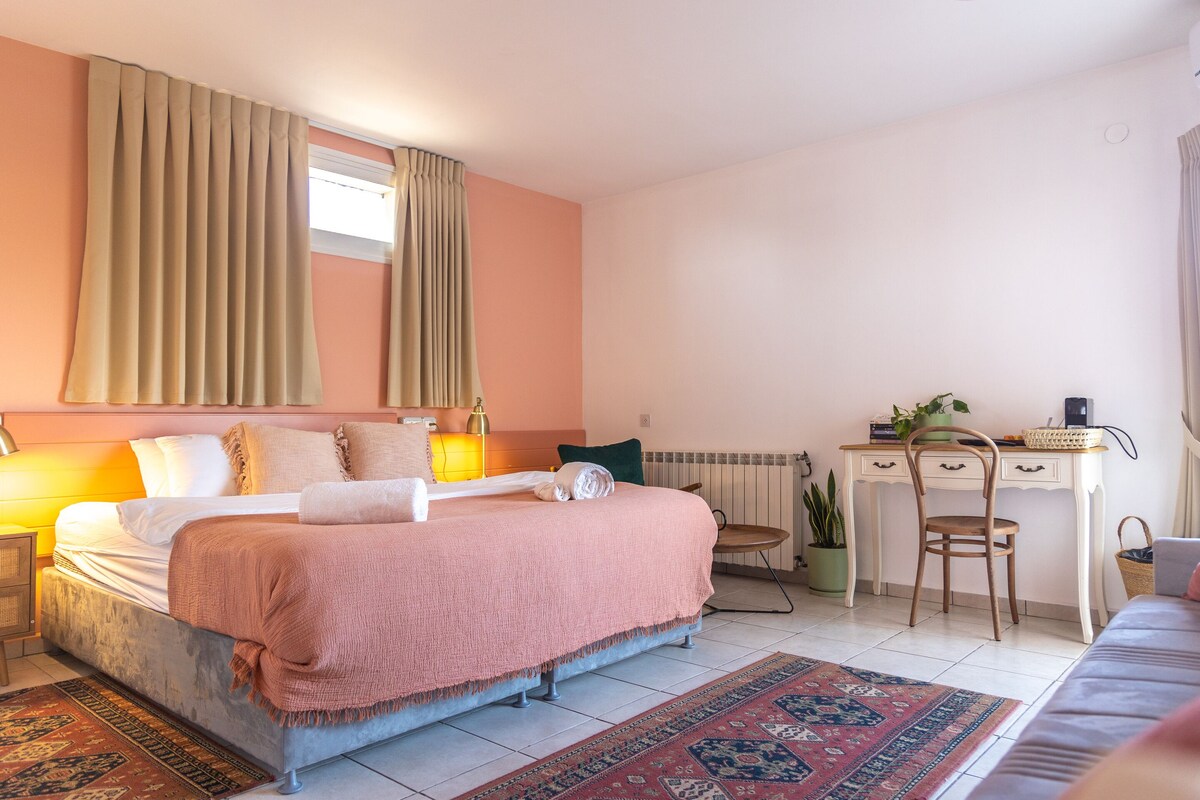 A spacious bedroom features a large bed with light linens and a coral-colored throw. Warm lighting is provided by bedside lamps, and sheer curtains allow natural light. A desk and chair sit near a potted plant, while a decorative rug adds warmth to the tiled floor.
