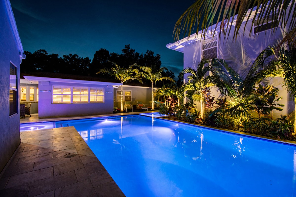 The illuminated salt-water pool glows in vibrant blue under a night sky, surrounded by tropical landscaping and palm trees. The nearby patio area features outdoor seating and the home is visible in the background, bathed in soft lighting that enhances the peaceful atmosphere.