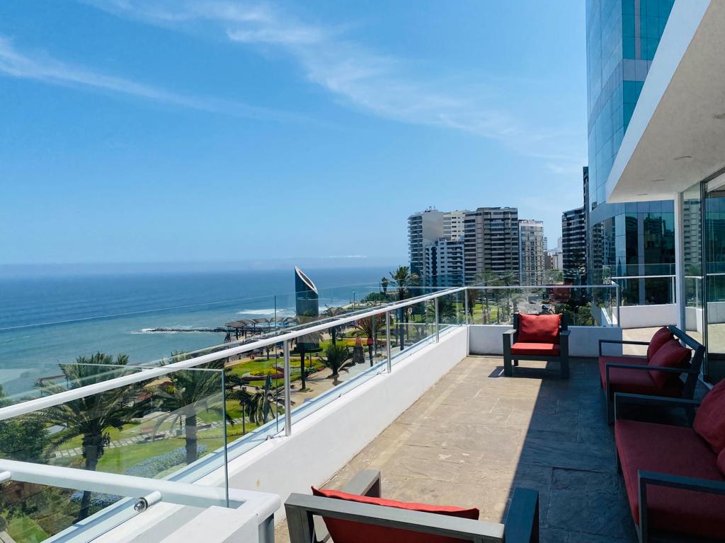 A spacious terrace features a modern seating area, with comfortable red chairs positioned along a glass railing. A clear view of the ocean and nearby skyline is visible, creating a relaxed outdoor space for enjoying the scenery. Lush greenery can be seen in the distance.