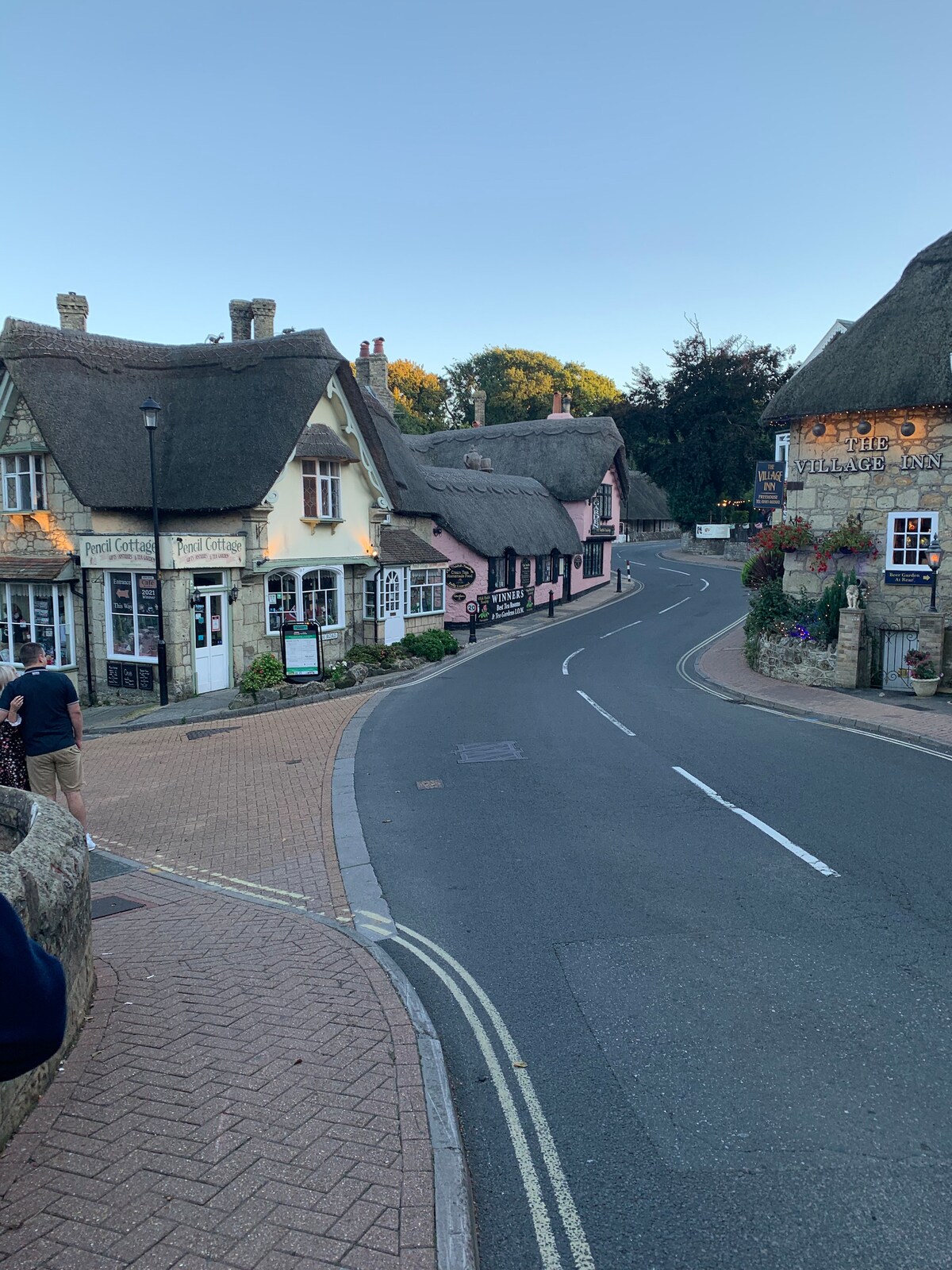 A view of Shanklin Old Village showcases charming thatched-roof cottages lining a gently curving street. The cobblestone road is flanked by vibrant floral displays and quaint signage. A clear sky is visible, adding to the peaceful ambiance of the setting.