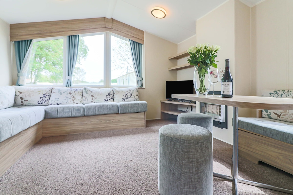 A welcoming living area is showcased, featuring large windows that allow natural light to fill the space. A comfortable corner sofa with decorative cushions is present alongside a small dining table set with a flower vase and a bottle. Neutral tones create a relaxing environment.