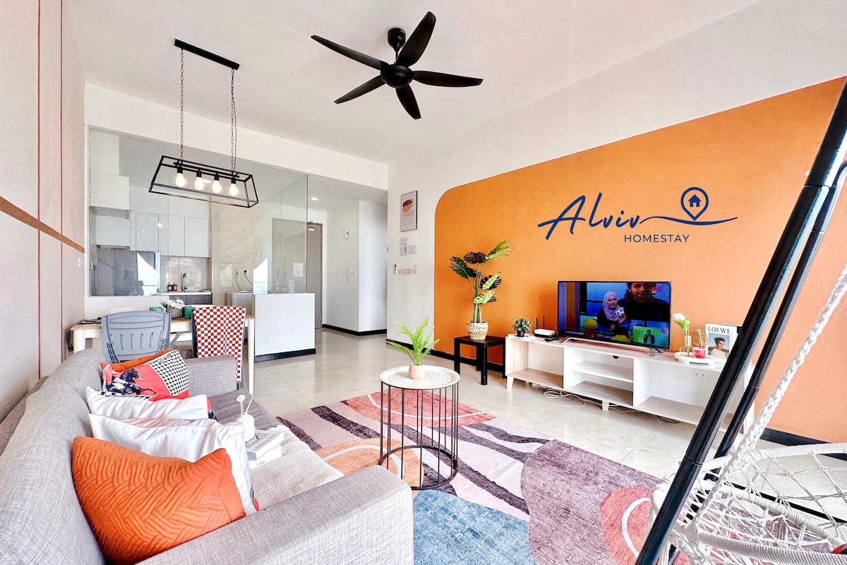 A spacious living area is showcased with a comfortable sofa adorned with decorative pillows. A circular table sits center stage, alongside a TV mounted on a stand with entertainment options displayed. Bright orange walls contrast with the light-colored furniture, while a ceiling fan provides airflow.