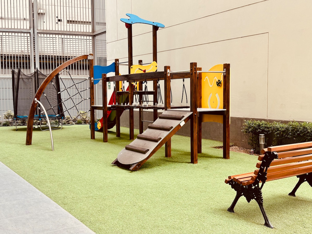 A colorful children's playground features a climbing structure, slide, and activity panels. A green synthetic grass surface surrounds the play area, providing a safe space for children. A wooden bench is positioned nearby for parents to relax while supervising their children.