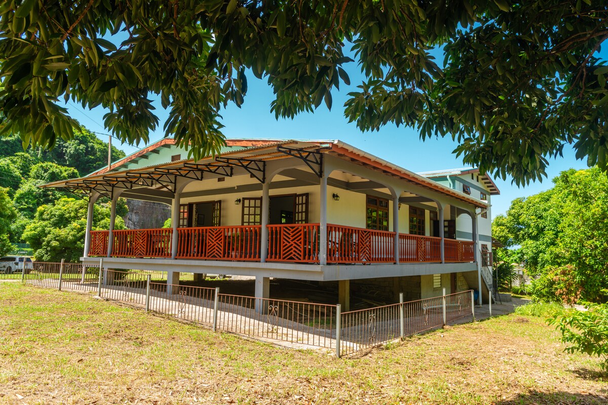A spacious house is set in a lush garden, surrounded by trees. The property features a large verandah with wooden railings that offer shaded outdoor space. The house is elevated, allowing for an expansive view of the green surroundings and natural landscape.