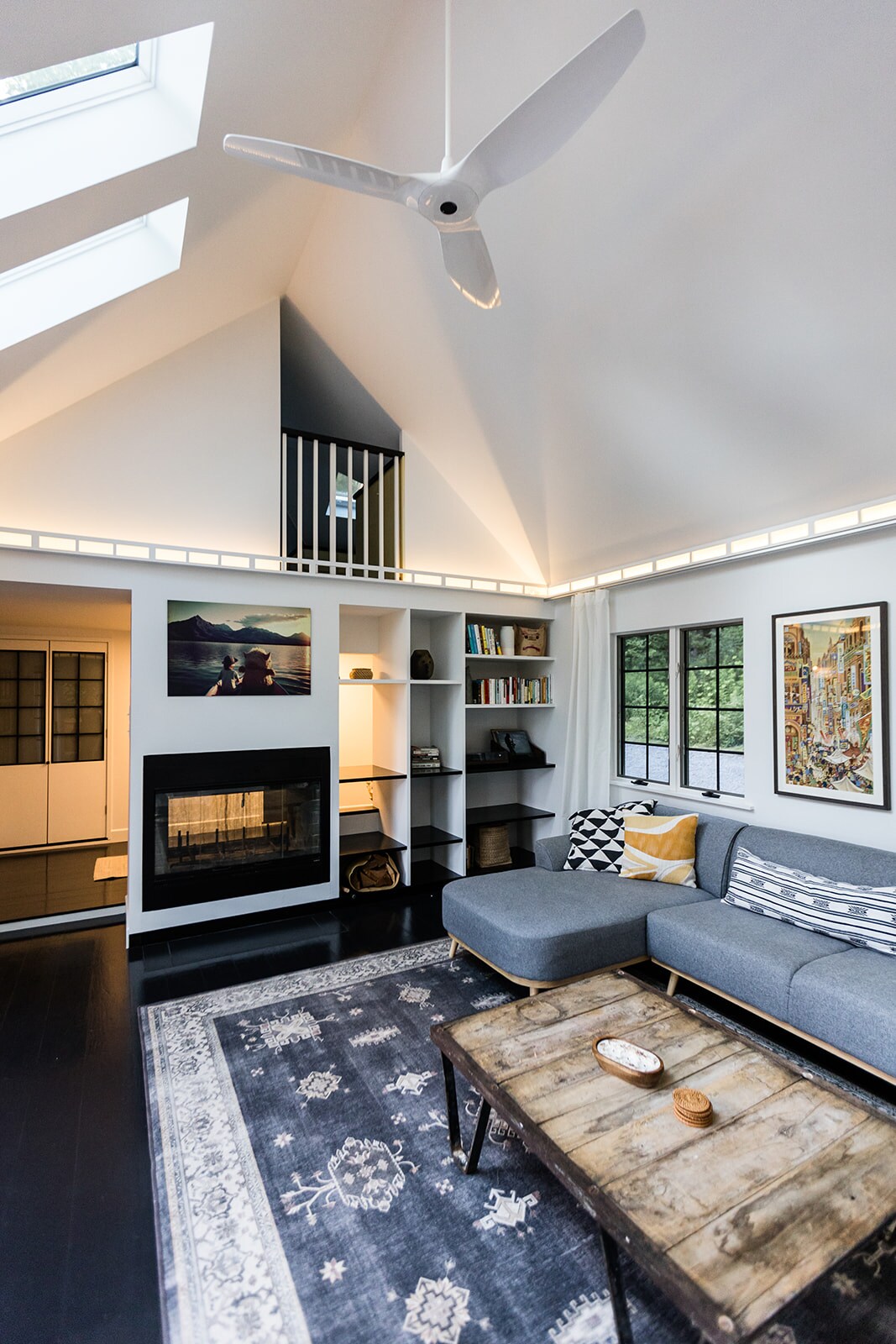 A modern living space is illuminated by natural light from large skylights and windows. A cozy sectional sofa faces a rustic coffee table situated on a patterned rug. A mounted fan and a contemporary electric fireplace enhance the area, while bookshelves and artwork add character.