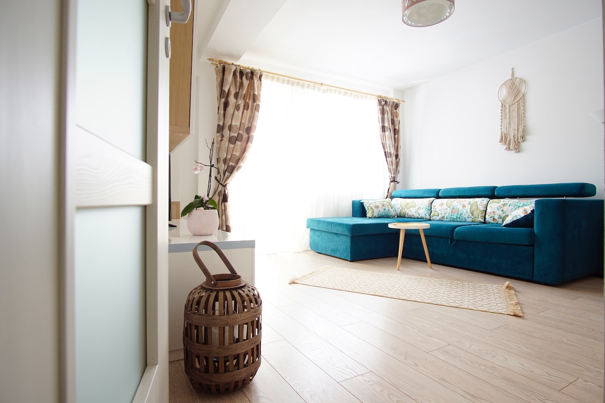A bright living area is presented, featuring a comfortable teal sofa adorned with floral cushions. A small wooden table sits on a light rug, while natural light filters through sheer curtains, enhancing the spacious feel of the room. A decorative basket and plant add subtle touches.