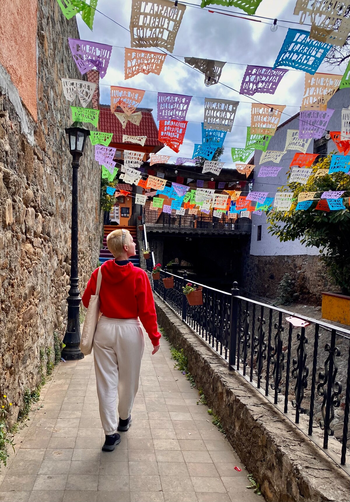 A narrow pathway is framed by rustic stone walls and colorful papel picado banners overhead. A person walks away along the cobblestone path, leading towards a glimpse of a tranquil waterway. The scene captures the vibrant local culture and serene atmosphere.