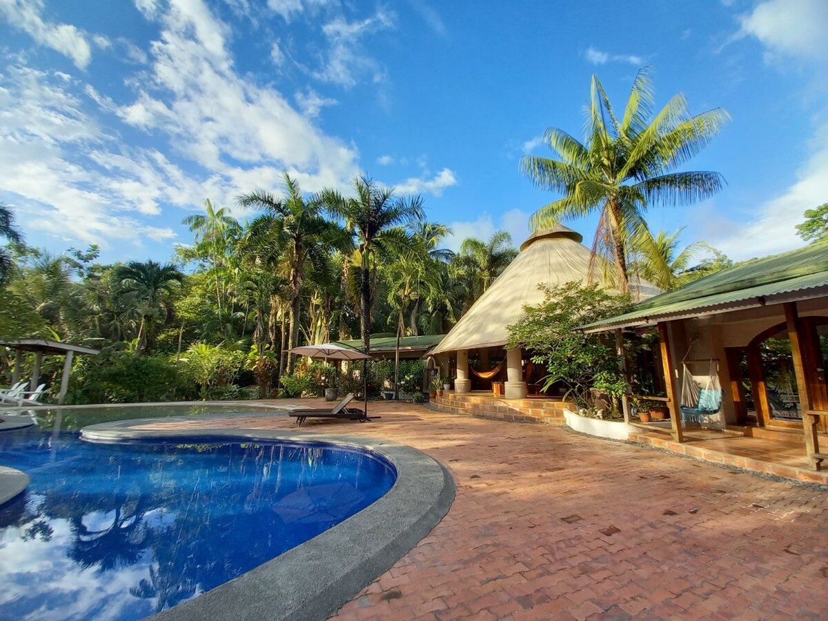 A round, thatched-roof structure is situated beside a sparkling swimming pool, framed by tropical palm trees. The spacious patio hosts lounge chairs and shaded umbrellas, inviting relaxation. Lush greenery surrounds the area, creating a serene atmosphere in this tranquil outdoor setting.