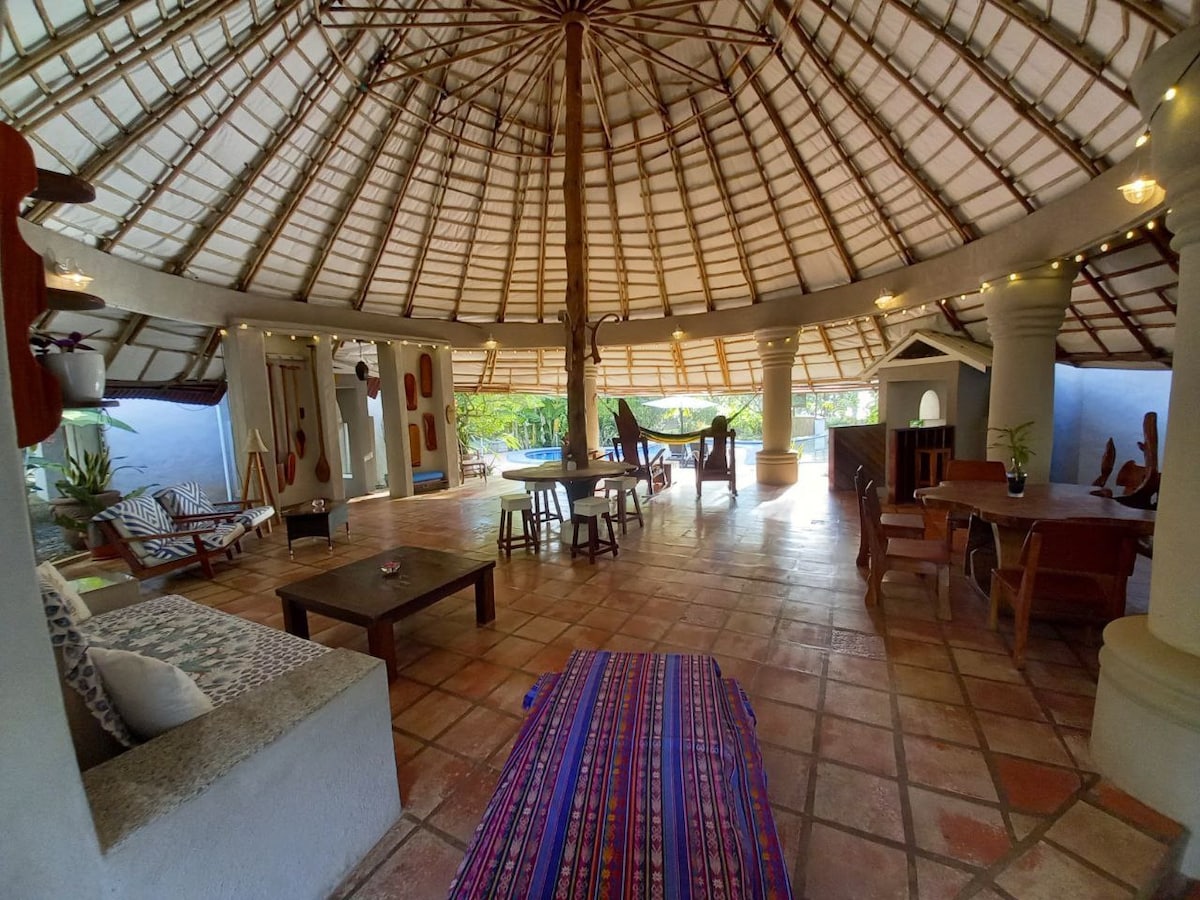 An open-plan living area features a high, circular thatched roof supported by a central pillar. Rustic wooden furniture, including tables and chairs, is arranged across terracotta tiles. Decorative elements and lush greenery are visible, contributing to a serene ambiance.