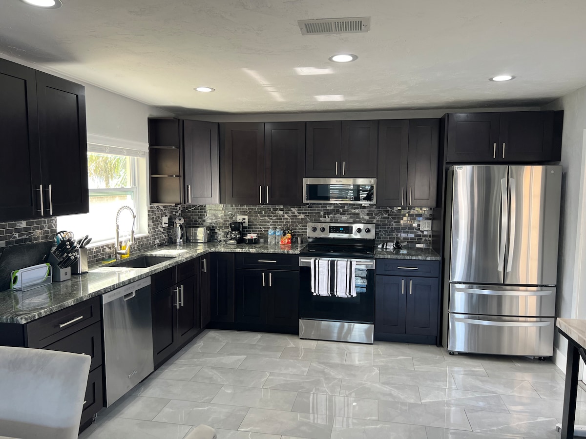 A modern kitchen displays sleek dark cabinetry and stainless steel appliances, including a refrigerator and oven. Light quartz countertops provide a functional workspace. Natural light fills the room through a window above the sink, highlighting the tiled backsplash and ample storage.