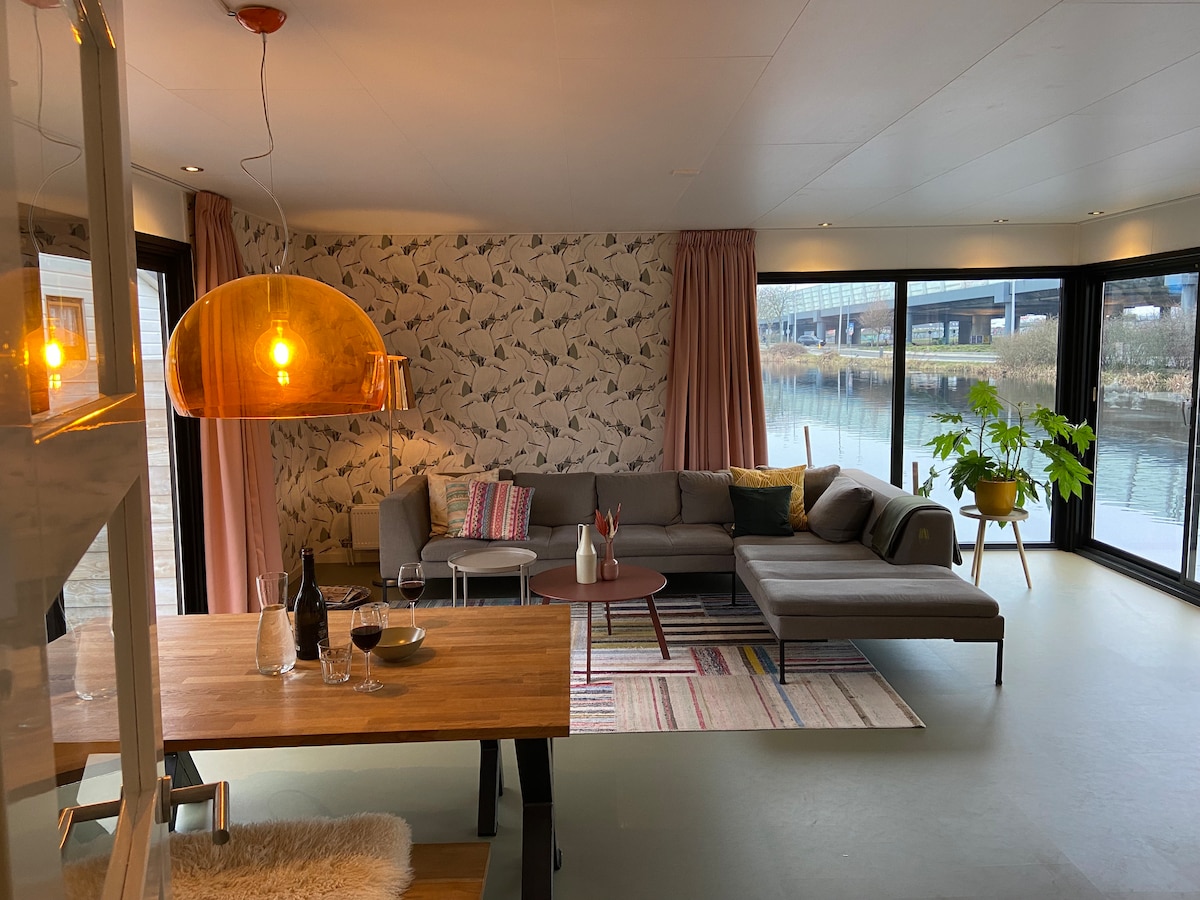 An open living space is presented with large windows that provide views of the water. A comfortable sectional sofa is paired with a round coffee table and a dining table set for four. Soft lighting is created by a pendant light, adding warmth to the space.
