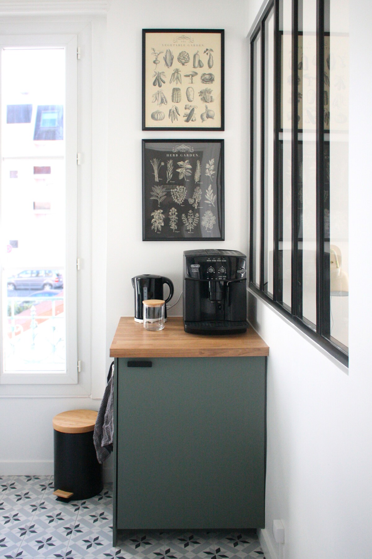 A cozy corner is highlighted with a wooden countertop hosting a coffee maker and glass of water. Decorative prints hang on the wall above, while a small stool and a grey cabinet complete the space. Natural light filters through the window nearby.