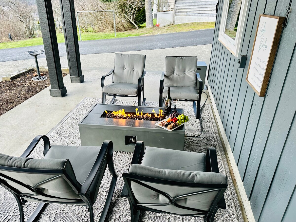 An outdoor seating area features four plush chairs arranged around a modern fire table, which displays a flickering flame. A subtle area rug enhances the setting, complemented by a nearby charcoal barbecue and a scenic view of the surrounding landscape.