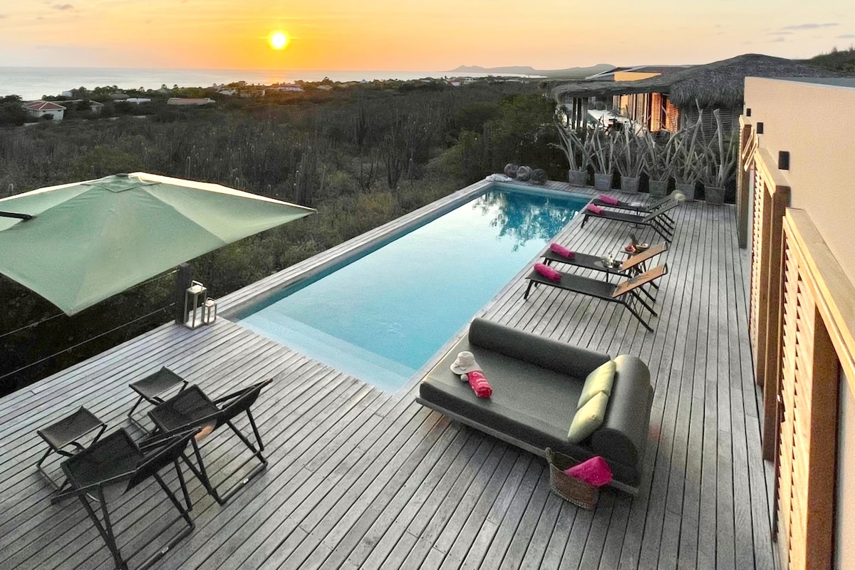 A spacious wooden deck surrounds a modern swimming pool, with sun loungers arranged thoughtfully and an umbrella providing shade. The sunset casts warm hues across the sky, highlighting the serene coastal landscape and distant hills.