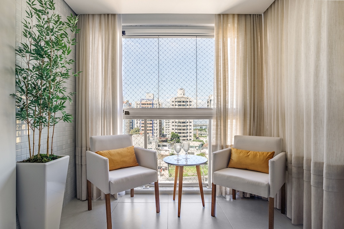 A bright sitting area features two light-colored armchairs with orange cushions positioned around a small table. Natural light filters through sheer curtains, offering a view of the cityscape beyond a mesh-screened window. A potted plant adds a touch of greenery to the space.