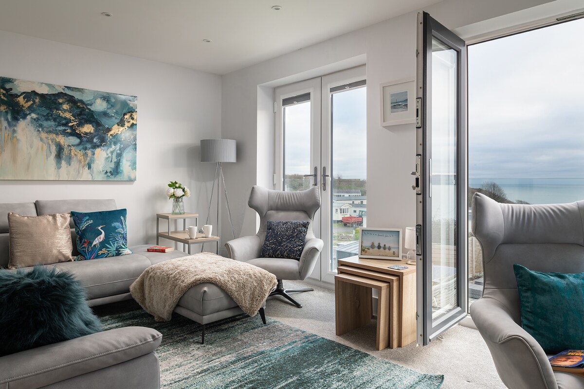 A modern living area showcases comfortable seating, with two armchairs positioned beside a stylish couch. Natural light floods the space through large glass doors, which open to a stunning sea view. An artistic wall hanging adds a vibrant touch to the neutral decor.