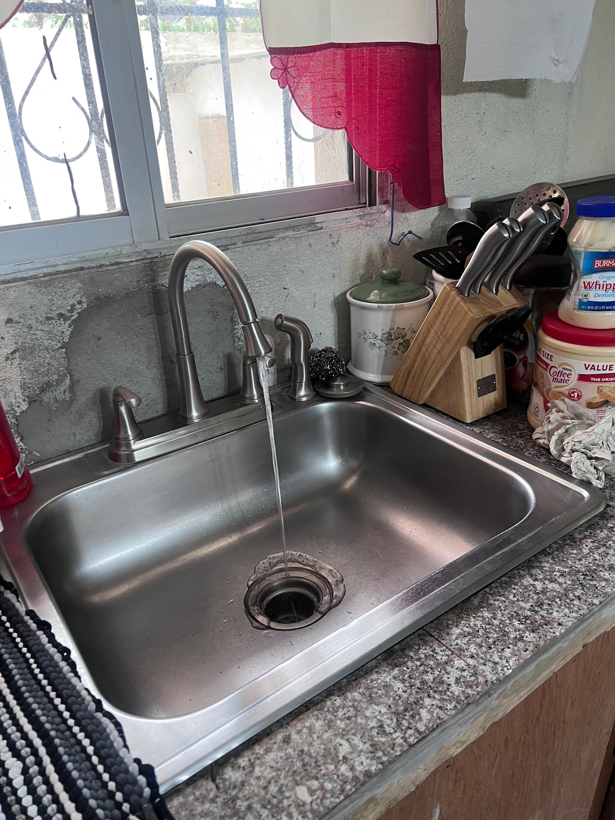 A stainless steel sink is displayed with water flowing from the faucet. Nearby, various kitchen utensils are arranged, including a knife block and containers for cooking ingredients, all positioned against a granite countertop with a window providing natural light.