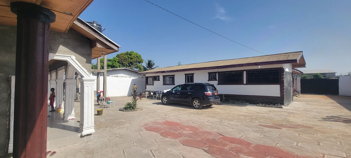 An outdoor view of the property reveals a spacious driveway lined with patterned tiles. A dark-colored vehicle is parked beside a light-colored building featuring multiple windows. Lush greenery is present, enhancing the environment's peacefulness.