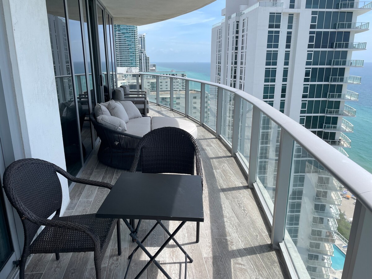 A wrap-around balcony is showcased, featuring a small black table and two wicker chairs. The ocean can be seen in the distance, accompanied by the skyline of nearby buildings. Natural light brightens the space, creating a relaxing outdoor retreat.