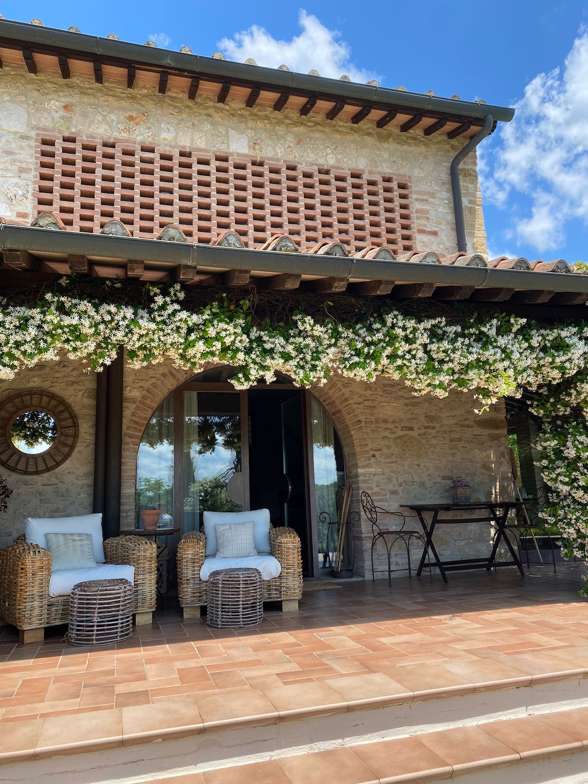 An exterior view of the dwelling features a spacious terrace adorned with flowering vines. Two wicker chairs with soft cushions and a small table create a comfortable seating area. Large glass doors provide access to the interior, framed by stone walls and a tiled floor.