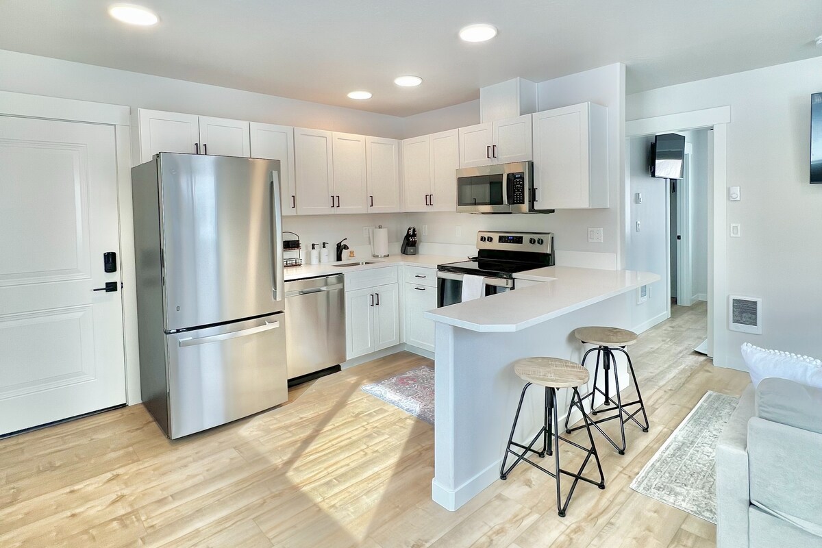 A modern kitchen is shown, featuring stainless steel appliances and white cabinetry. A spacious island is accompanied by two tall stools, and natural light fills the space through overhead lighting. A living area is visible in the background, enhancing the open layout.