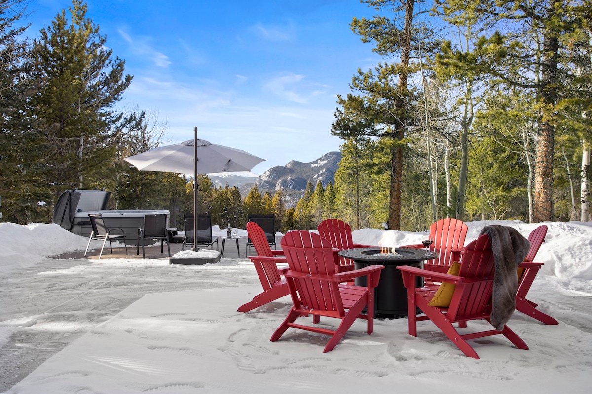 A fire pit area features red Adirondack chairs arranged around a central fire pit, set against a backdrop of snow-covered ground and tall evergreen trees. A patio umbrella provides shade, and distant mountains are visible under a clear blue sky.