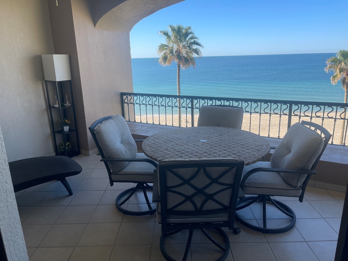 A private patio features a round table surrounded by four cushioned swivel chairs. A view of the ocean and sandy beach is visible beyond the railing, complemented by palm trees swaying gently in the breeze under a clear blue sky.