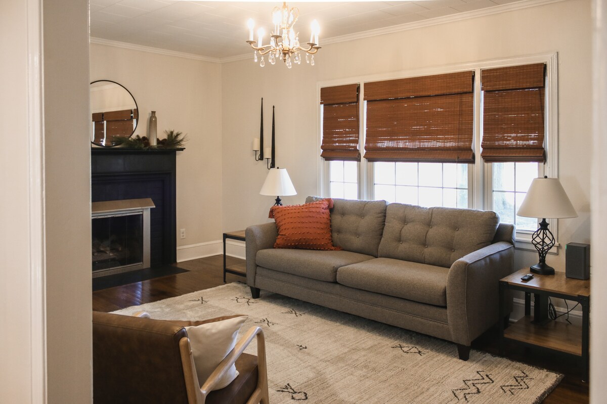 A living room is shown featuring a gray fabric couch adorned with an orange accent pillow. A gas fireplace is positioned under a round mirror, with two lamps illuminating the space. Natural light enters through the windows, complemented by bamboo shades.
