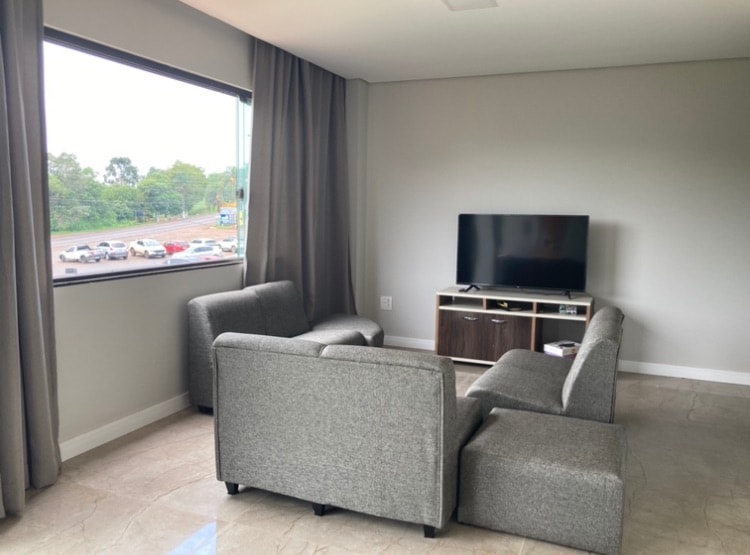 A seating area features two gray sofas arranged around a coffee table, with a flat-screen television mounted on a light brown media console. A large window offers a view of the outside space, allowing natural light to enter the room.