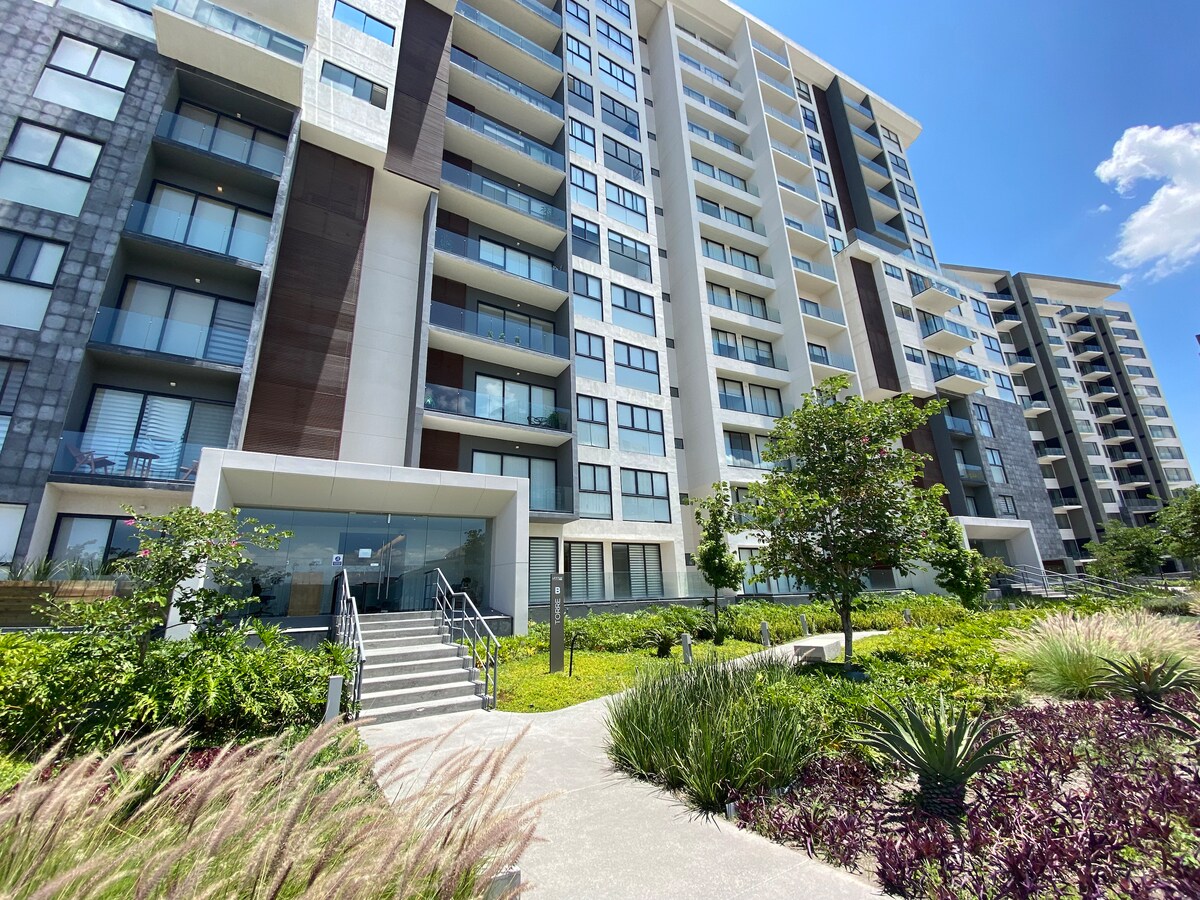 The exterior of the apartment building showcases a modern design with a mixture of materials. Lush greenery surrounds the entrance, featuring landscaped areas with various plants and pathways leading up to the main entrance, which is framed by large glass panels.