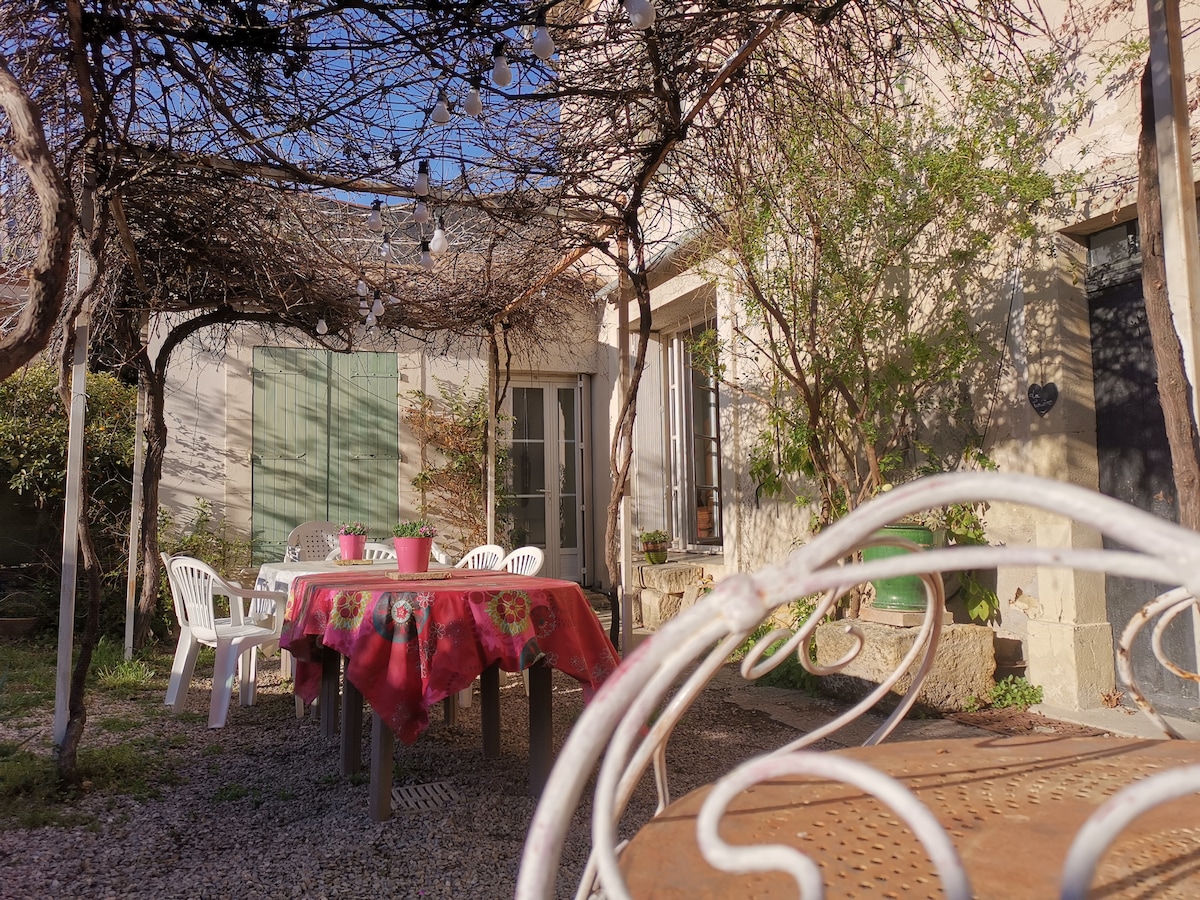 An outdoor dining area is visible under a vine-covered trellis, featuring a large table adorned with a colorful tablecloth and surrounded by white chairs. The sun-soaked garden setting enhances the inviting atmosphere of this peaceful retreat.