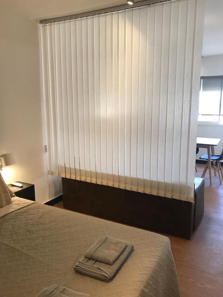 A well-organized sleeping area is defined by vertical blinds that create a separation from the adjacent space. The bed is neatly made, with towels placed at the foot. A simple table with chairs is visible in the background, complemented by natural light from the window.