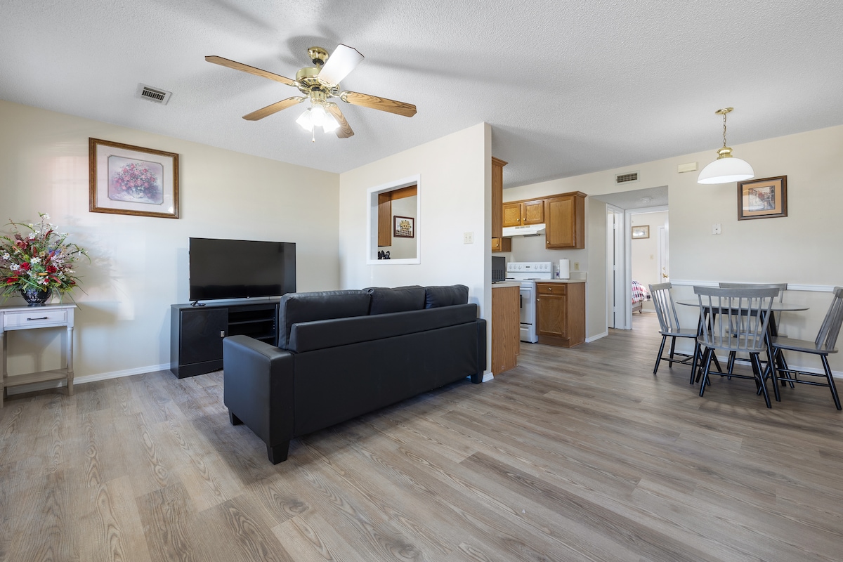 A living area is furnished with a dark sofa and a television on a stand. A ceiling fan circulates air, and natural light enters through windows. The open layout leads to a dining area with a table and four chairs, adjacent to a kitchen.