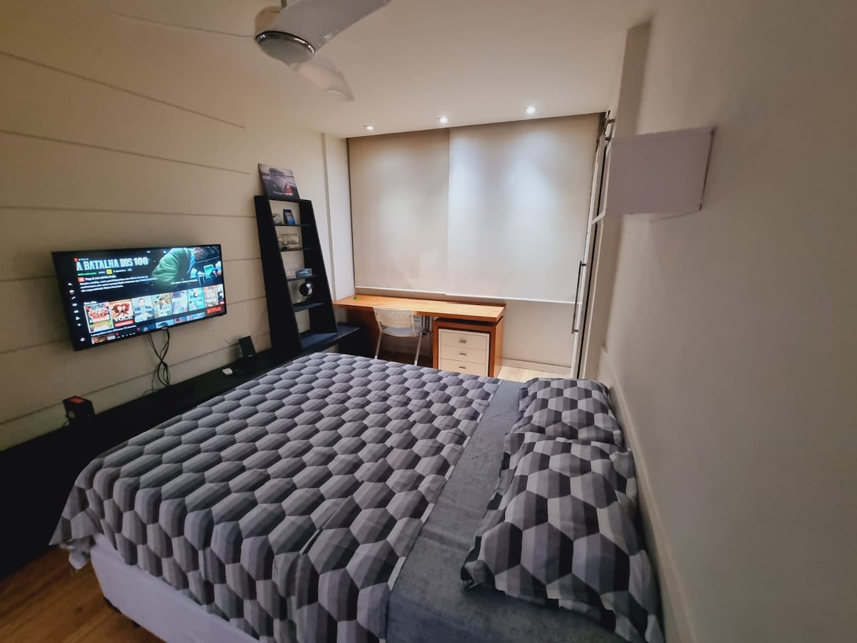 A comfortable bedroom is showcased with a spacious bed featuring a geometric patterned cover. A TV mounted on the wall offers entertainment options. A study area with a desk and chair is illuminated by soft overhead lighting, creating a functional space.