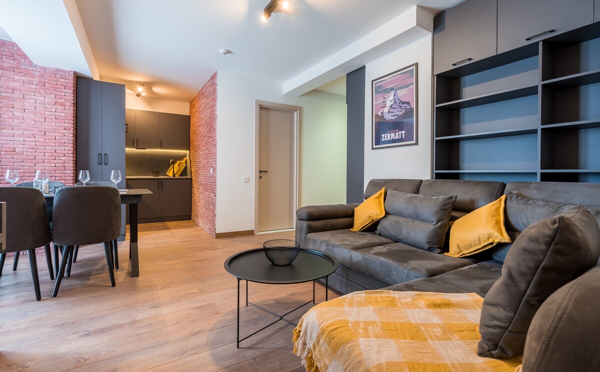 The living area features a comfortable sofa with decorative cushions, a round coffee table, and a neatly arranged dining space. A modern kitchenette with dark cabinetry is visible in the background, along with a striking brick accent wall and a framed artwork.