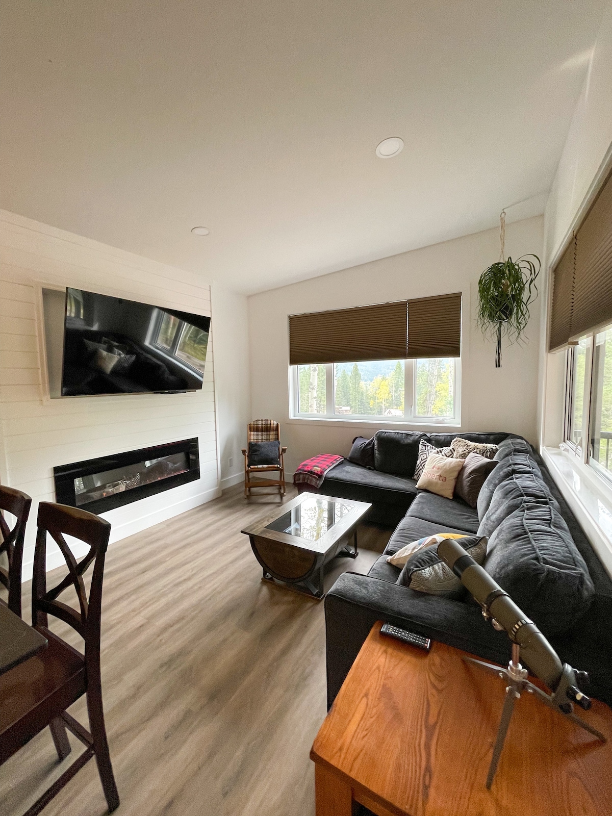 A cozy living space features a modern sectional couch facing a wall-mounted television. A glass coffee table sits in front, while a wooden side table with a remote is nearby. Large windows with brown shades offer natural light, complemented by a mounted planter.