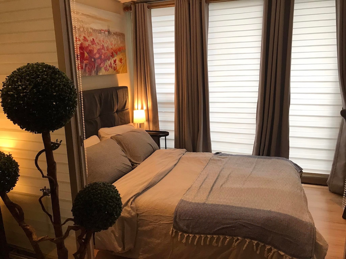 A cozy sleeping area is highlighted by a spacious bed with soft grey bedding. Two tall windows are dressed with sheer curtains, allowing natural light to filter in. A decorative plant stands nearby, complementing the warm ambiance created by a bedside lamp.