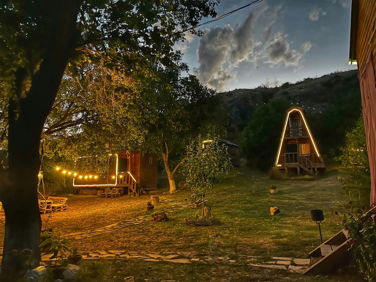 An outdoor area is illuminated by string lights, surrounded by trees and a mountain backdrop. A triangular-shaped cabin is positioned next to a wooden deck, with grassy lawns providing ample space for relaxation. Soft twilight enhances the tranquil environment.
