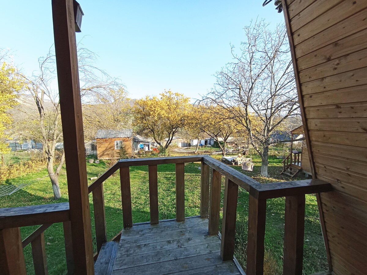 A wooden balcony provides a view of a grassy yard dotted with yellow-leaved trees. Small structures are visible in the distance, blending into a serene natural landscape under a clear blue sky.