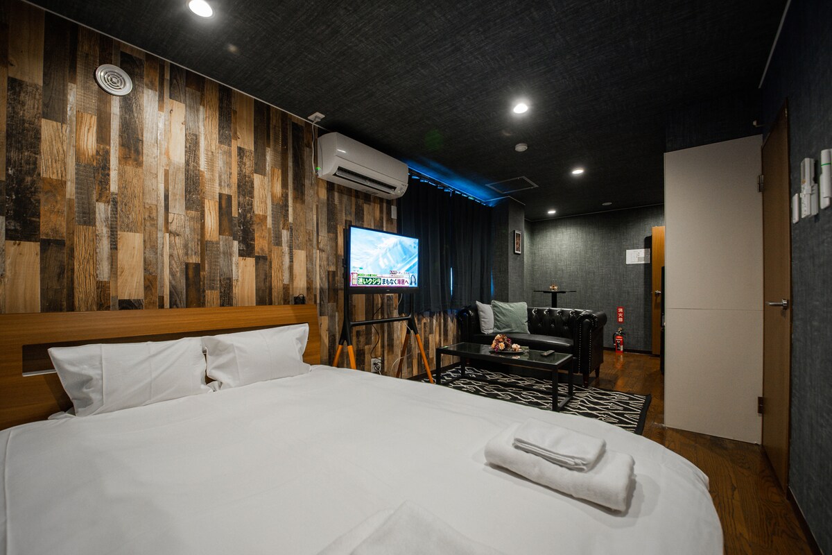 A spacious room features a large bed with crisp white linens and a cozy seating area. A black leather sofa complements the wooden accents on the walls. A mounted television is positioned across from the bed, providing entertainment options.