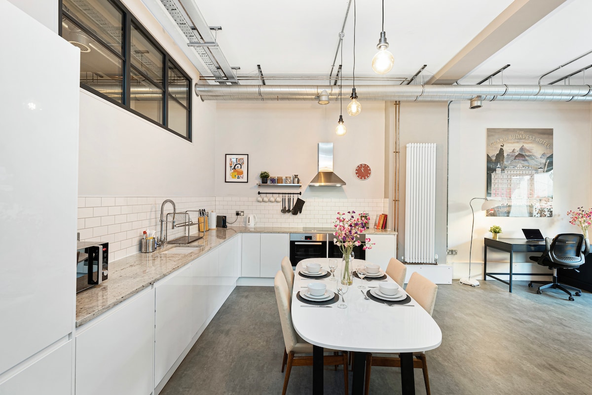 A modern kitchen area features sleek cabinetry and a spacious countertop. A dining table is set with plates and flowers, while appliances, including a coffee maker and oven, are visible. Track lighting and large windows provide a bright, inviting space, enhancing its contemporary design.