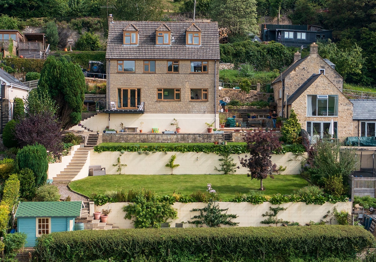 A detached house is situated overlooking a landscaped tiered garden, which features lawn areas and several planting beds. Steps lead from the terrace to the garden. The structure includes multiple windows, providing ample natural light, while the surrounding greenery enhances the tranquil setting.