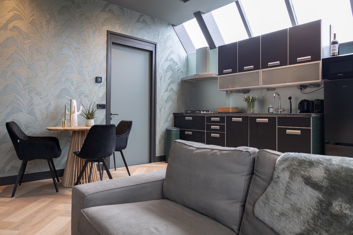 A modern kitchen area features sleek cabinetry and integrated appliances. A dining table with two chairs is set beside the window, while a comfortable sofa offers a cozy seating option. Natural light floods the space through multiple skylights, highlighting the wallpaper's botanical design.