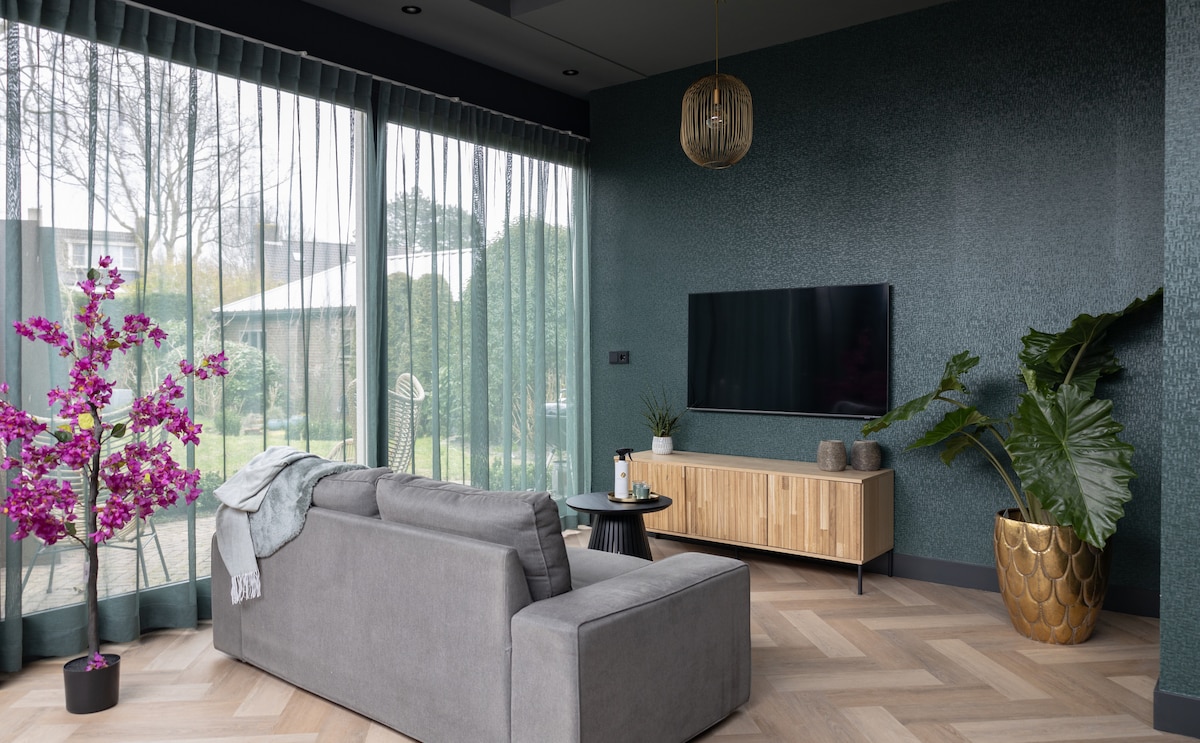 A tastefully furnished living area is highlighted by a plush gray sofa facing a mounted television. Sheer green curtains gently filter light, revealing a well-tended garden outside. A decorative plant and a flowering pink tree add vibrant touches to the serene atmosphere.