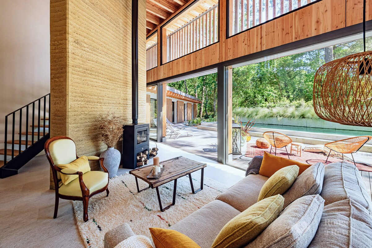 A spacious living area features large glass doors that open to a lush outdoor space, allowing natural light to fill the room. A comfortable sofa is positioned in front of a modern fireplace, with a wooden coffee table and decorative items on a soft area rug.