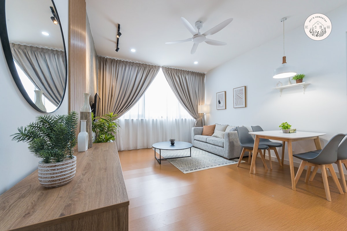 A modern living space is furnished with a plush sofa, a circular coffee table, and a dining area featuring a light wood table with four chairs. Natural light filters through floor-to-ceiling curtained windows, complemented by indoor plants and minimalist decor.