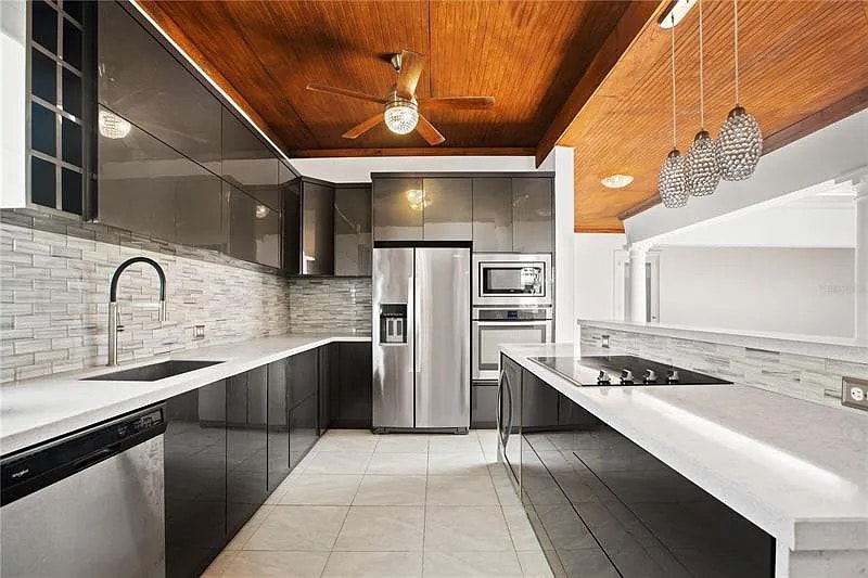 The modern kitchen features sleek cabinetry and a spacious layout. Stainless steel appliances, including a refrigerator, oven, and dishwasher, are complemented by a marble countertop. Natural light enters through large windows, creating a bright cooking space with a ceiling fan overhead.